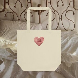 Pode incluir: Bolsa tote creme com um gráfico de coração rosa que diz "LOVE YOURS". A bolsa tem duas alças e é feita de um tecido resistente. A bolsa tote é usada para transportar itens.