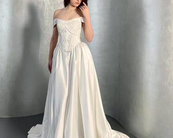 Könnte beinhalten: Ein schulterfreies, bodenlanges Kleid mit einem weißen Oberteil mit aufwendiger Blumenstickerei und einer strukturierten Korsett-Taille. Der Rock besteht aus glattem, einfarbigem weißem Satin, der in eine volle, voluminöse Silhouette fällt.