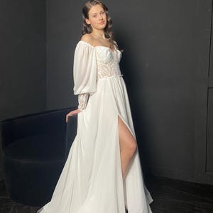 May include: A white off-the-shoulder wedding dress with a high slit. The bodice features intricate lace detailing, and the long sleeves have a puffy design. The dress has a flowing skirt and a train.