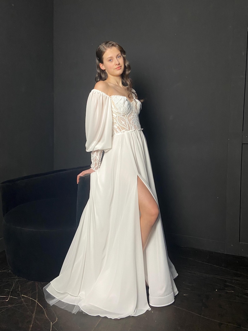 May include: A white off-the-shoulder wedding dress with a high slit. The bodice features intricate lace detailing, and the long sleeves have a puffy design. The dress has a flowing skirt and a train.