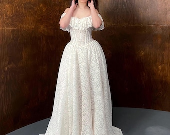 May include: A floor-length white lace gown featuring an off-the-shoulder neckline with a ruffled collar, a fitted corset-style bodice with vertical boning, and a full, voluminous skirt made of intricate floral lace fabric.
