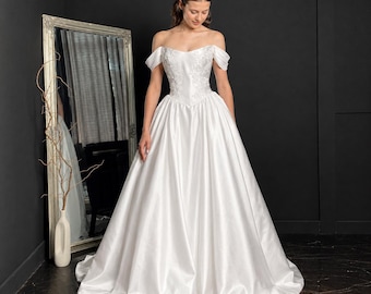 Könnte beinhalten: Ein bodenlanges weißes Brautkleid mit schulterfreiem Ausschnitt, einem eng anliegenden Oberteil mit detaillierter weißer Blumenstickerei und einem voluminösen Satinrock, der von der Taille bis zum Boden in einer weiten Form fällt.