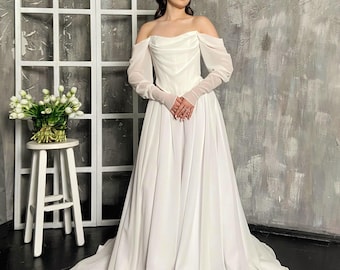 May include: A white off-the-shoulder wedding dress with long, sheer sleeves. The gown features a fitted bodice and a flowing skirt. The model is wearing sheer gloves. A bouquet of white tulips sits on a small white table.