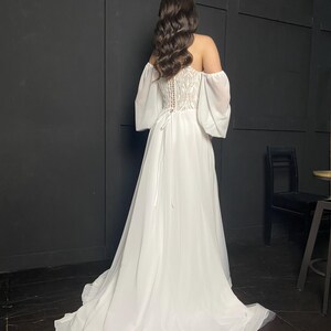 May include: A white off-the-shoulder wedding dress with a flowing skirt and a detailed bodice. The dress features sheer sleeves and a lace-up back. The model's dark hair is styled in loose waves.