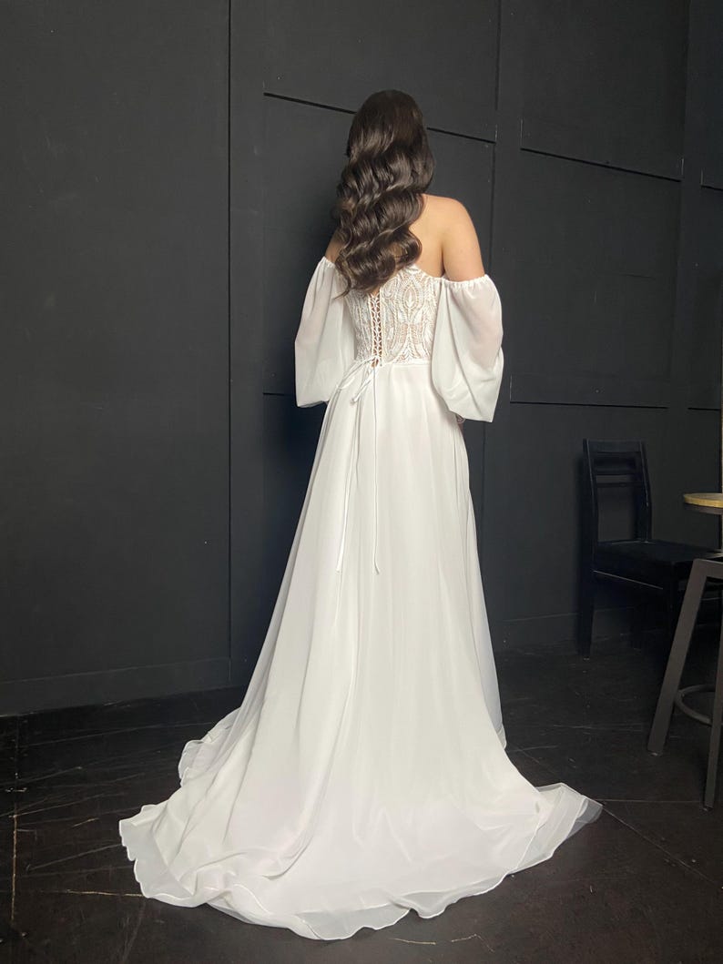 May include: A white off-the-shoulder wedding dress with a flowing skirt and a detailed bodice. The dress features sheer sleeves and a lace-up back. The model's dark hair is styled in loose waves.