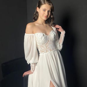 May include: White off-the-shoulder wedding dress with a lace bodice and long, sheer sleeves. The dress features a high slit and a flowing skirt. The model is smiling, with dark hair and a black background.