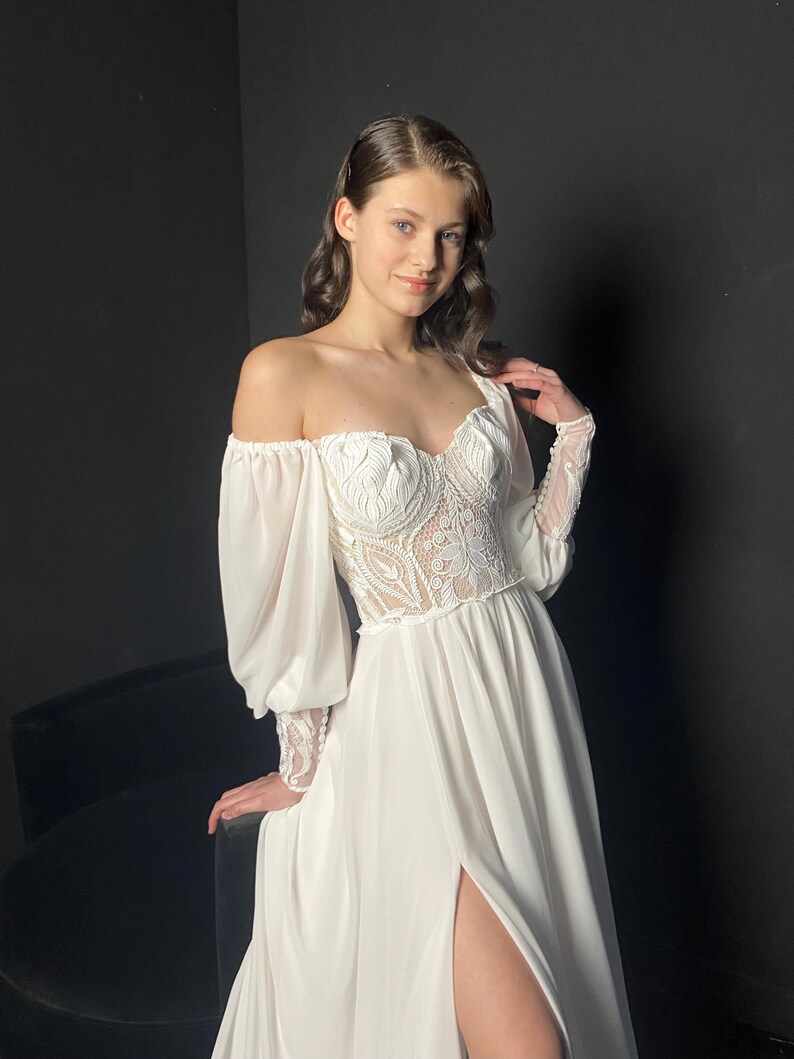 May include: White off-the-shoulder wedding dress with a lace bodice and long, sheer sleeves. The dress features a high slit and a flowing skirt. The model is smiling, with dark hair and a black background.