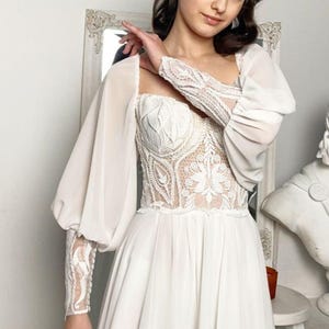 May include: A white wedding dress with a square neckline and long, sheer sleeves. The bodice features intricate lace detailing, and the skirt flows to the floor. The dress has a high slit, and the overall style is elegant and romantic.