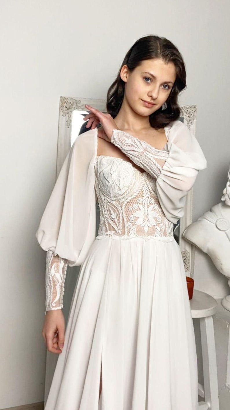 May include: A white wedding dress with a square neckline and long, sheer sleeves. The bodice features intricate lace detailing, and the skirt flows to the floor. The dress has a high slit, and the overall style is elegant and romantic.