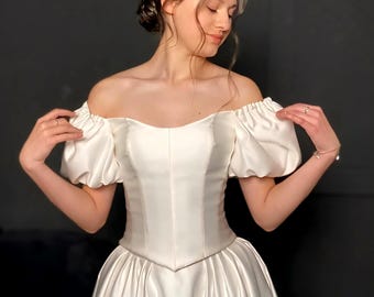 May include: A white off-the-shoulder dress with short, puffed sleeves. The bodice is fitted, and the skirt flows gracefully. The dress appears to be made of a satin-like material, creating a smooth, elegant look. The overall style suggests a formal or bridal gown.