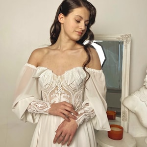 May include: An off-the-shoulder white wedding dress with a detailed lace bodice and long, sheer sleeves. The dress features a flowing skirt and is designed with intricate floral patterns.
