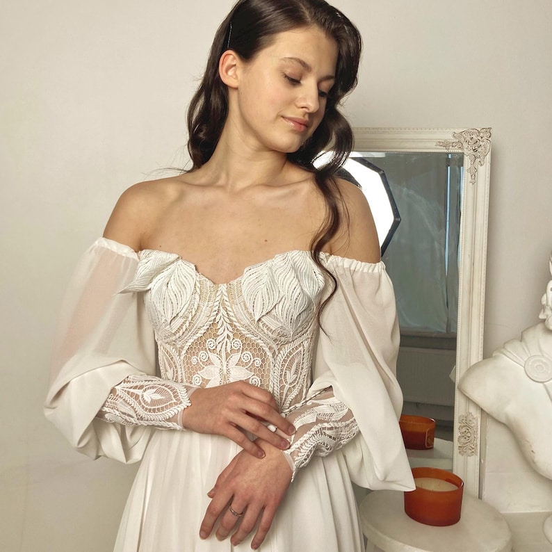 May include: An off-the-shoulder white wedding dress with a detailed lace bodice and long, sheer sleeves. The dress features a flowing skirt and is designed with intricate floral patterns.
