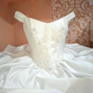 May include: A white satin wedding dress with an off-the-shoulder neckline. The bodice is adorned with delicate floral lace appliqués and sparkling embellishments. The skirt is full and flowing, creating a classic bridal silhouette.