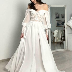 May include: A white off-the-shoulder wedding dress with a fitted bodice and flowing skirt. The bodice features intricate lace detailing, and the long sleeves are sheer. The dress has a romantic, ethereal feel, perfect for a special occasion.
