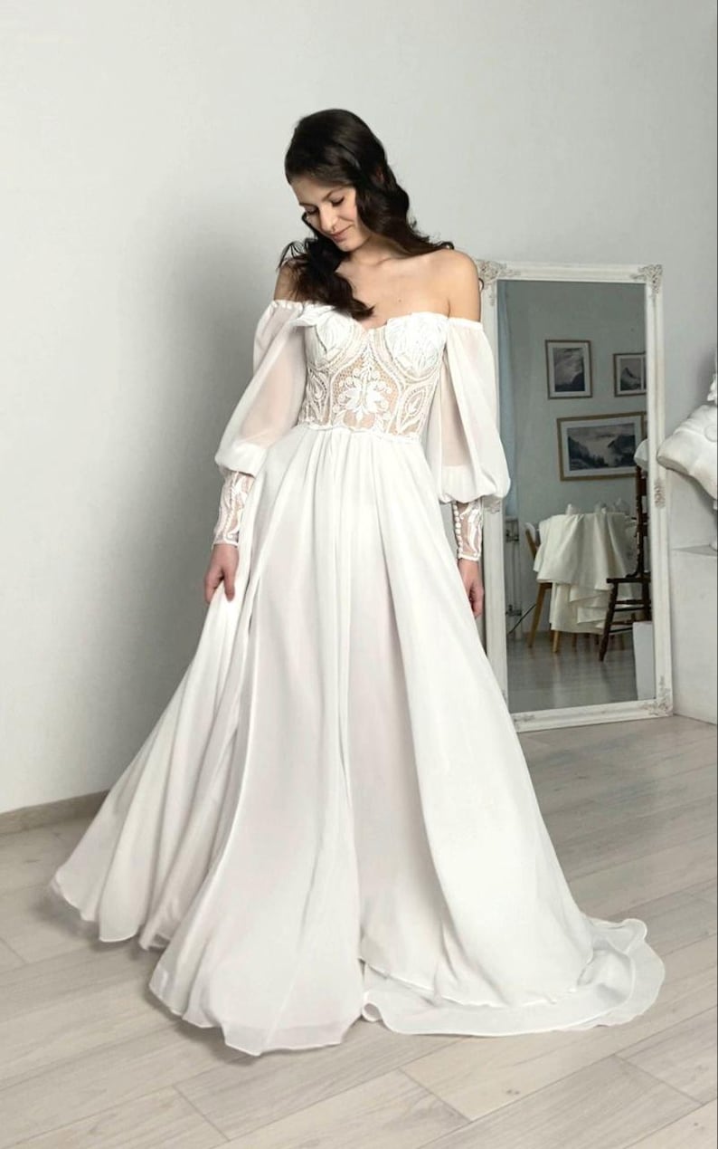 May include: A white off-the-shoulder wedding dress with a fitted bodice and flowing skirt. The bodice features intricate lace detailing, and the long sleeves are sheer. The dress has a romantic, ethereal feel, perfect for a special occasion.