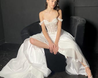 May include: A white off-the-shoulder gown featuring a structured corset bodice with intricate floral lace detailing and a voluminous, layered floor-length skirt. The garment is paired with white pointed-toe heels with a single ankle strap.