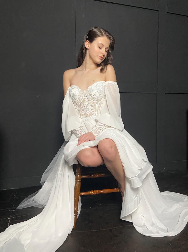 May include: A white off-the-shoulder wedding dress with a detailed bodice and flowing skirt. The dress features long, sheer sleeves and a train. The model is seated on a wooden chair.