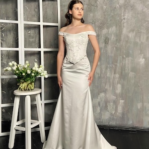 May include: A white off-the-shoulder wedding dress with a fitted bodice and a flowing skirt. The bodice features intricate embroidery. The dress is floor-length and has a slight train. The background is a neutral gray.