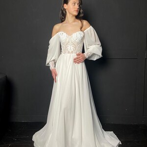 May include: A white off-the-shoulder wedding dress with a sweetheart neckline and long, flowing skirt. The bodice features intricate lace detailing, and the sleeves are long and sheer. The dress is floor-length and has a train.