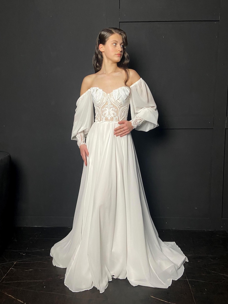 May include: A white off-the-shoulder wedding dress with a sweetheart neckline and long, flowing skirt. The bodice features intricate lace detailing, and the sleeves are long and sheer. The dress is floor-length and has a train.