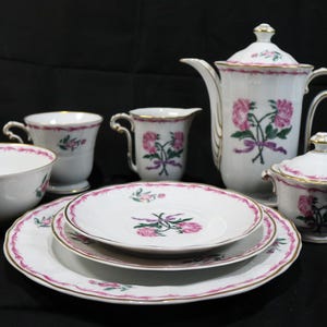 May include: A white porcelain tea set with pink floral designs and gold trim. The set includes a teapot, sugar bowl, creamer, cups, saucers, and a bowl. The floral pattern features pink roses and green leaves.