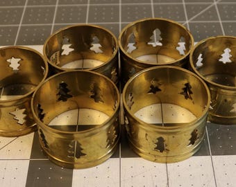 Könnte beinhalten: Sechs Serviettenringe aus Messing mit einem Weihnachtsbaum-Ausschnitt. Die Ringe sind zylindrisch und haben eine glänzende, goldene Oberfläche. Sie sind auf einer karierten Oberfläche angeordnet, was auf ein festliches oder weihnachtliches Thema hindeutet.