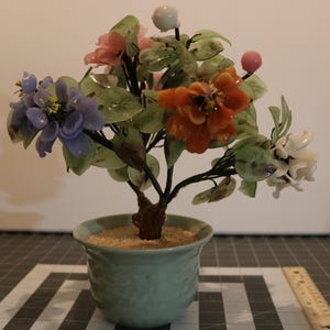 May include: A glass flower arrangement in a green ceramic pot. The flowers are made of jade and feature pink, orange, white, and purple blossoms.