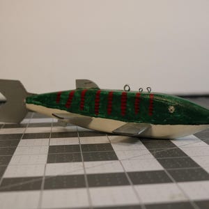 May include: A wooden fish lure painted green with red stripes. The lure has a silver tail fin and a white belly.