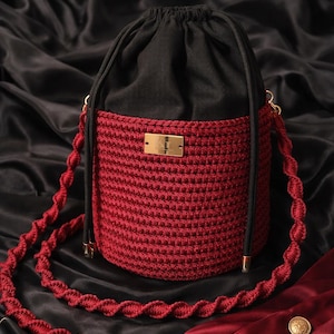 May include: A handmade red crochet bucket bag with a black drawstring top. The bag features a gold-tone metal label and a matching red, twisted strap. The bag is displayed against a black satin background.