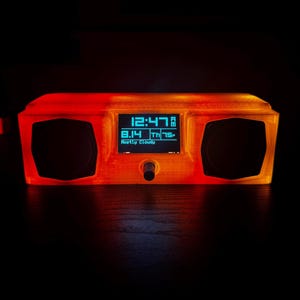 May include: A retro-style digital clock with a rectangular, orange-tinted casing. The clock face displays the time, date, and weather conditions. Two circular speaker openings flank the display. A small knob is centered below the screen.
