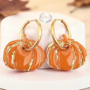 Può includere: Un paio di orecchini a cerchio dorati con ciondoli a forma di zucca arancione e dettagli in foglia d'oro. Ogni zucca è appesa a un cerchio dorato lucido. Il logo Lohyna è visibile al centro sopra gli orecchini. Il design è simmetrico.