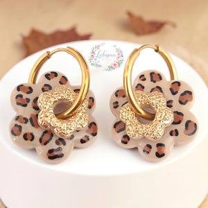 Può includere: Un paio di orecchini a cerchio color oro con un design a fiore a strati. Il fiore di base più grande ha una superficie beige con una stampa leopardata marrone e nera, mentre il fiore superiore più piccolo ha una finitura glitterata oro.