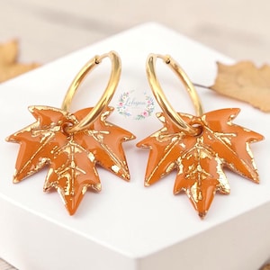 Può includere: Un paio di orecchini a cerchio dorati con pendenti a forma di foglia d'acero arancione, caratterizzati da venature e dettagli in oro metallizzato. Le foglie presentano una finitura liscia e lucida e sono appese a sottili cerchi dorati. Lohyna.