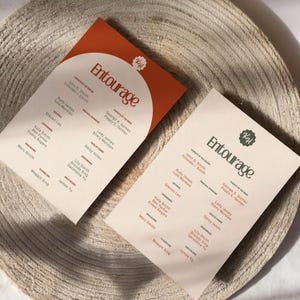 May include: Two beige cards with the word "Entourage" printed in brown text. The cards list names of people with titles like "Groom's Entourage" and "Bride's Entourage". The cards have a brown circle with the letter "KH" in white text.