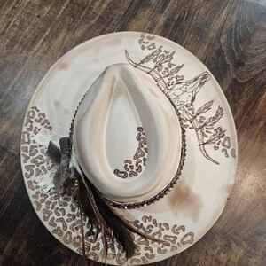 May include: A cream-colored cowboy hat with a wide brim featuring a brown leopard print pattern. The hat has a decorative band with sequins, feathers, and a brown steer skull design. The hat is a western-themed accessory.