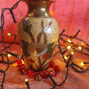 May include: A brass vase with a flared rim and a rounded body featuring a textured, cream-colored surface decorated with painted floral motifs in shades of purple, green, and red, surrounded by a string of small, glowing warm white lights.