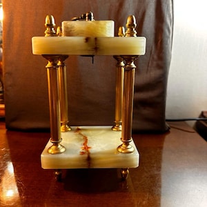 May include: A rectangular clock stand featuring two polished light green stone slabs with natural brown veining. Four vertical fluted brass pillars connect the top and bottom slabs, accented by small brass finials at the corners and four small feet.