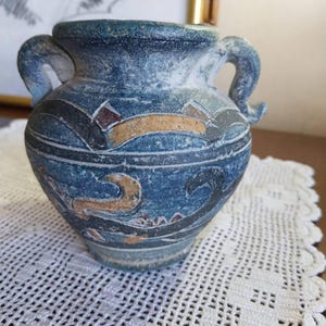 May include: A blue ceramic vase with two handles and decorative patterns in black, brown, and gold. The vase has a textured surface and a slightly weathered appearance, suggesting an antique or vintage style. The vase is sitting on a white lace doily.