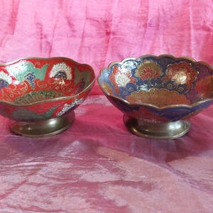May include: Two metal bowls with scalloped edges and pedestal bases. The left bowl features a red background with green, white, and gold floral patterns. The right bowl has a dark blue background with gold, red, and white floral patterns.