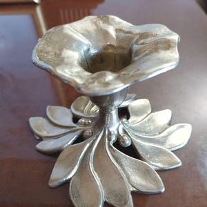 Peut inclure: Un bougeoir en métal argenté doté d'une base en forme de grappe de feuilles lisses et arrondies, surmonté d'une coupelle au bord évasé rappelant une fleur. La surface présente une finition polie et réfléchissante avec une texture subtile.
