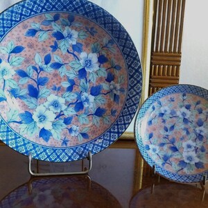 May include: Two round ceramic plates featuring a central design of blue and white flowers with green leaves set against a soft peach background. The rims are decorated with a dark blue and white geometric grid pattern, and both plates rest on stands.