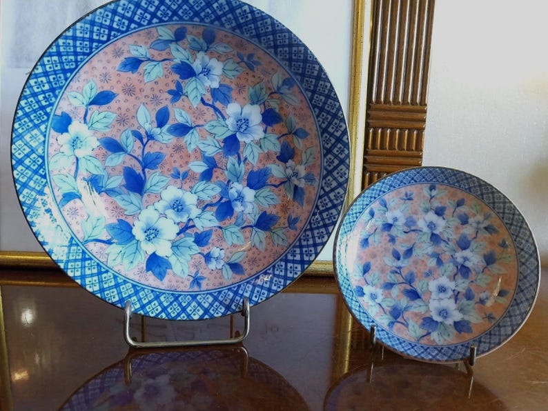 May include: Two round ceramic plates featuring a central design of blue and white flowers with green leaves set against a soft peach background. The rims are decorated with a dark blue and white geometric grid pattern, and both plates rest on stands.