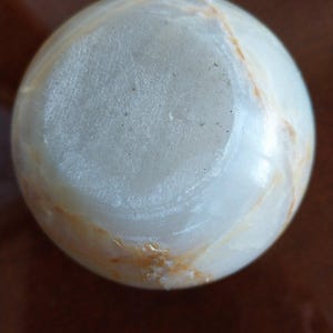 May include: A spherical object made of polished white stone with translucent qualities and visible natural veining in shades of tan and light brown. The surface features a smooth, rounded texture with a circular flat base area at the top.