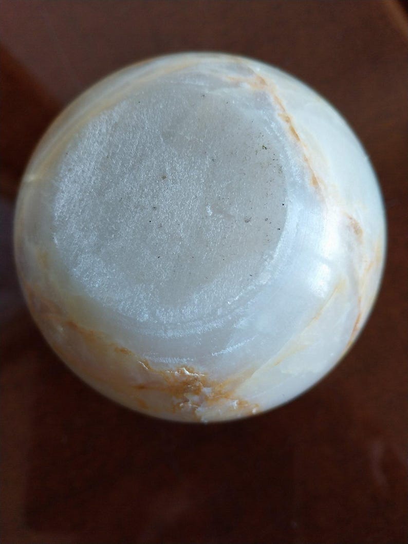 May include: A spherical object made of polished white stone with translucent qualities and visible natural veining in shades of tan and light brown. The surface features a smooth, rounded texture with a circular flat base area at the top.