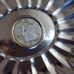 May include: A silver-toned metal serving dish with a fluted edge. The center features a circular design with two swans, water, and foliage. The dish has a shiny, reflective surface, and the design is detailed.