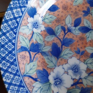 May include: A round ceramic plate featuring a central peach-colored background decorated with blue and white floral patterns and green leaves. The border displays a geometric blue and white grid design with small square accents throughout the pattern.