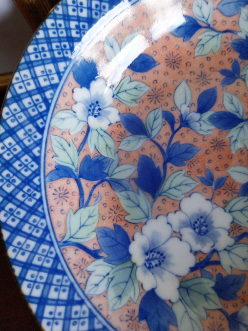 May include: A round ceramic plate featuring a central peach-colored background decorated with blue and white floral patterns and green leaves. The border displays a geometric blue and white grid design with small square accents throughout the pattern.
