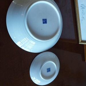 May include: Two white ceramic bowls shown from the underside, featuring a circular base and a subtle ribbed texture on the exterior. Each bowl has a small, square blue stamp with Asian characters centered on the bottom of the base.