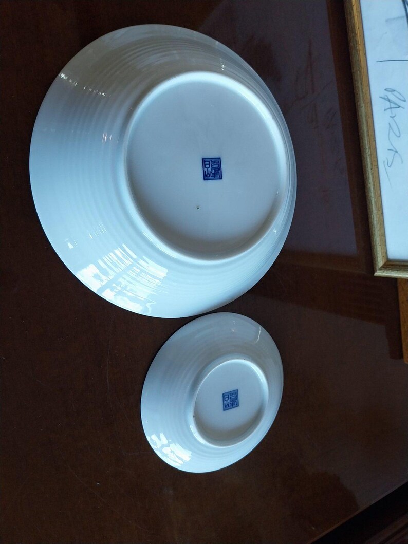 May include: Two white ceramic bowls shown from the underside, featuring a circular base and a subtle ribbed texture on the exterior. Each bowl has a small, square blue stamp with Asian characters centered on the bottom of the base.