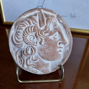 Puede incluir: Una escultura en relieve redonda de color terracota de un perfil clásico, montada sobre un pequeño soporte de metal. La escultura presenta una representación detallada del rostro de una persona con cabello rizado y un tocado decorativo. El relieve tiene detalles blancos.